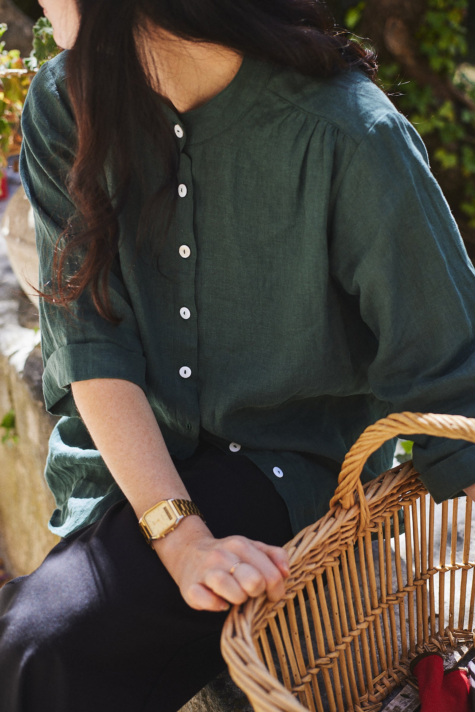 A person wearing a pine green blouse with mandarin style collar and 3/4 length sleeves, sitting next to a wicker basket.