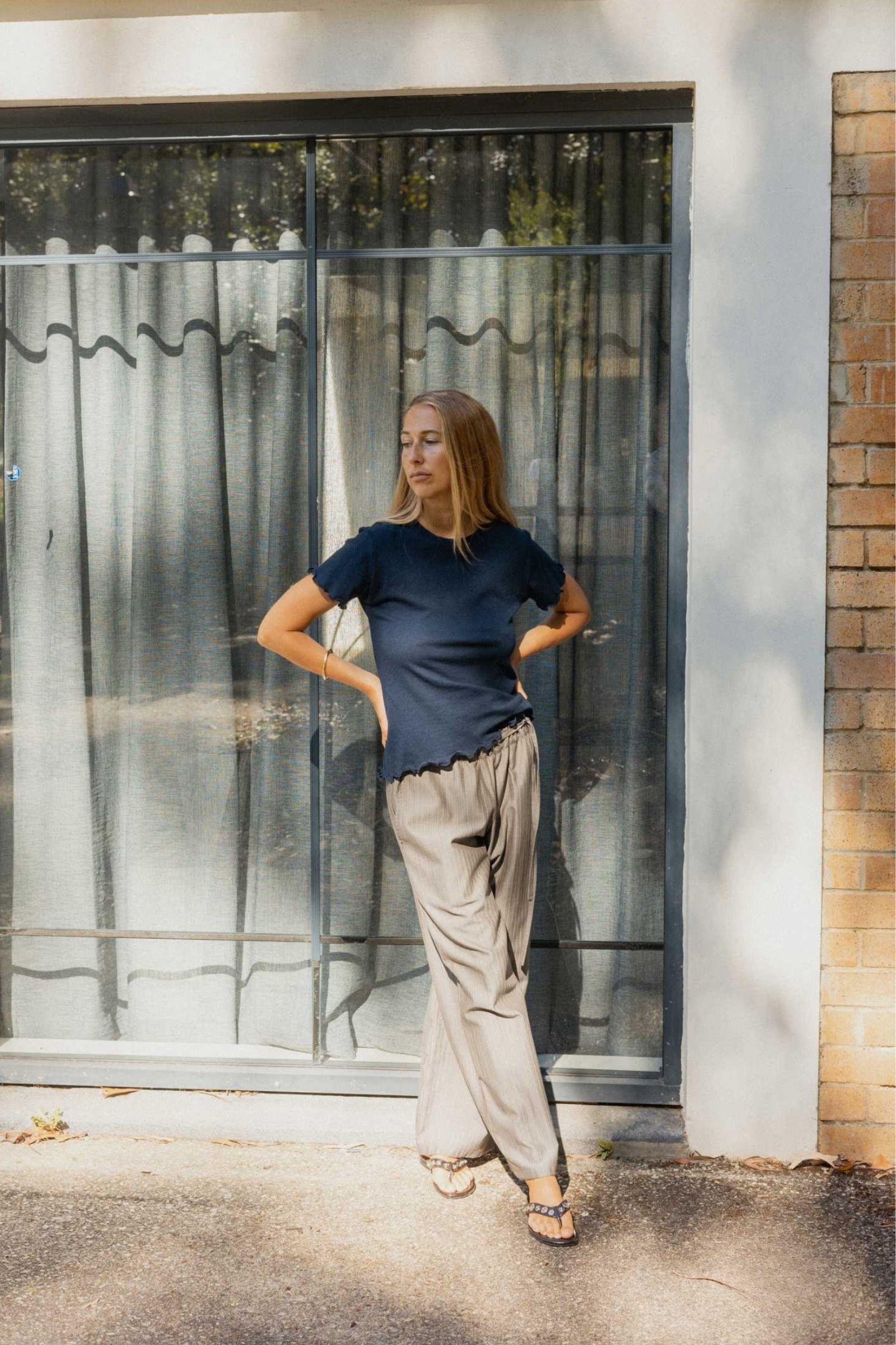 Woman standing in front of a glass door with metal bars, wearing a dark blue Tshirt and light-colored pants.