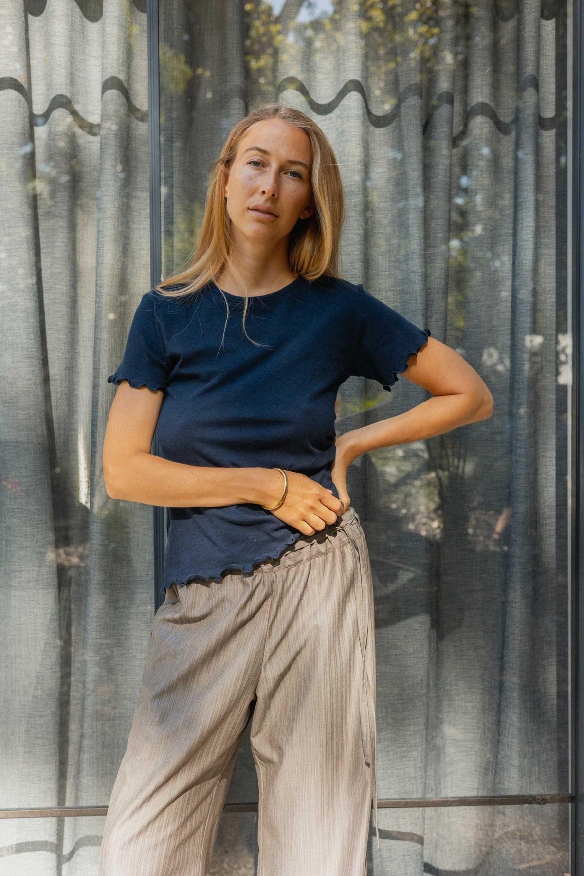 Woman wearing a navy blue tee and grey pants standing against a textured wall.