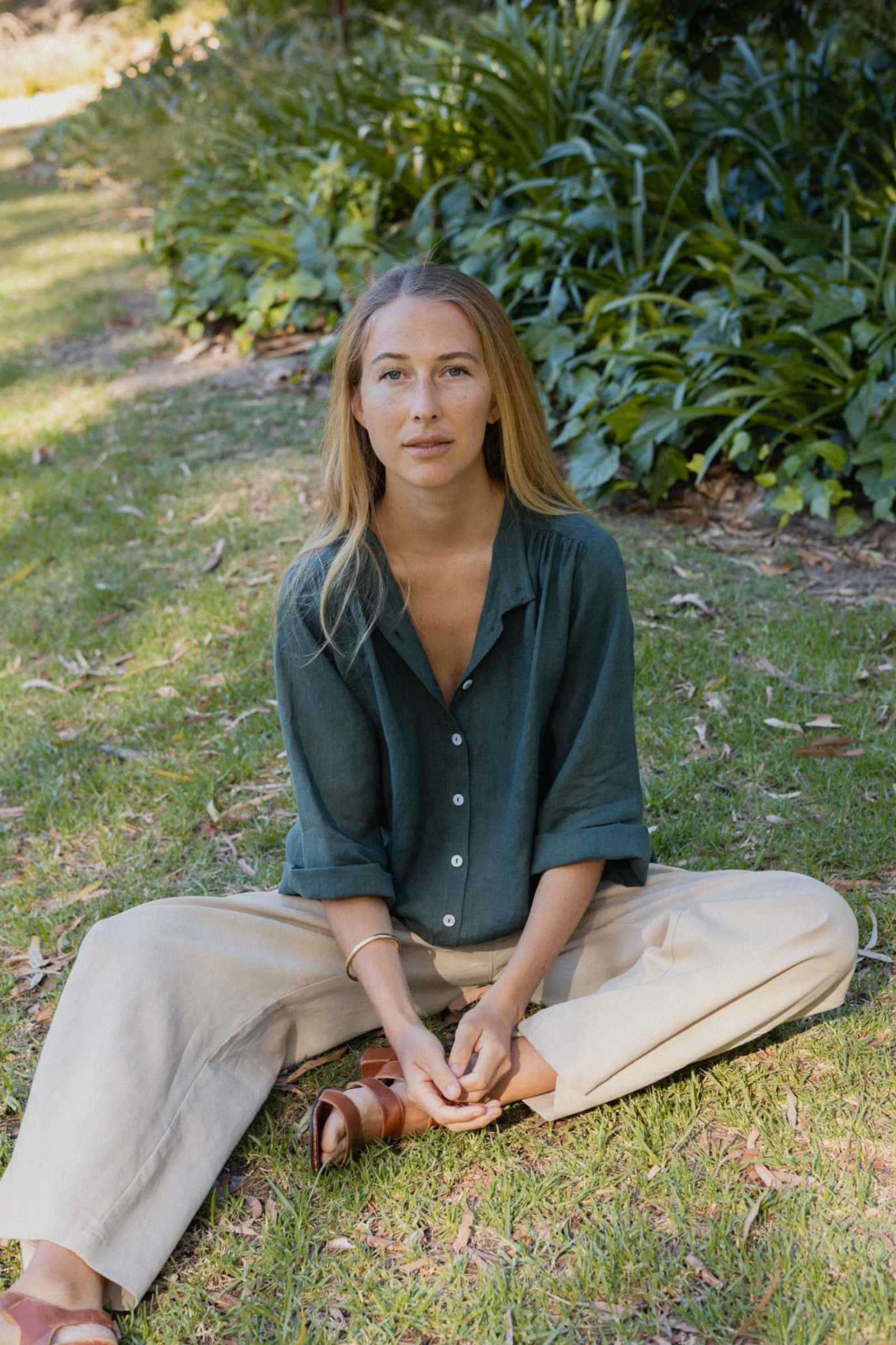 Woman sitting on grass wearing a green shirt and beige pants with a plant in the background