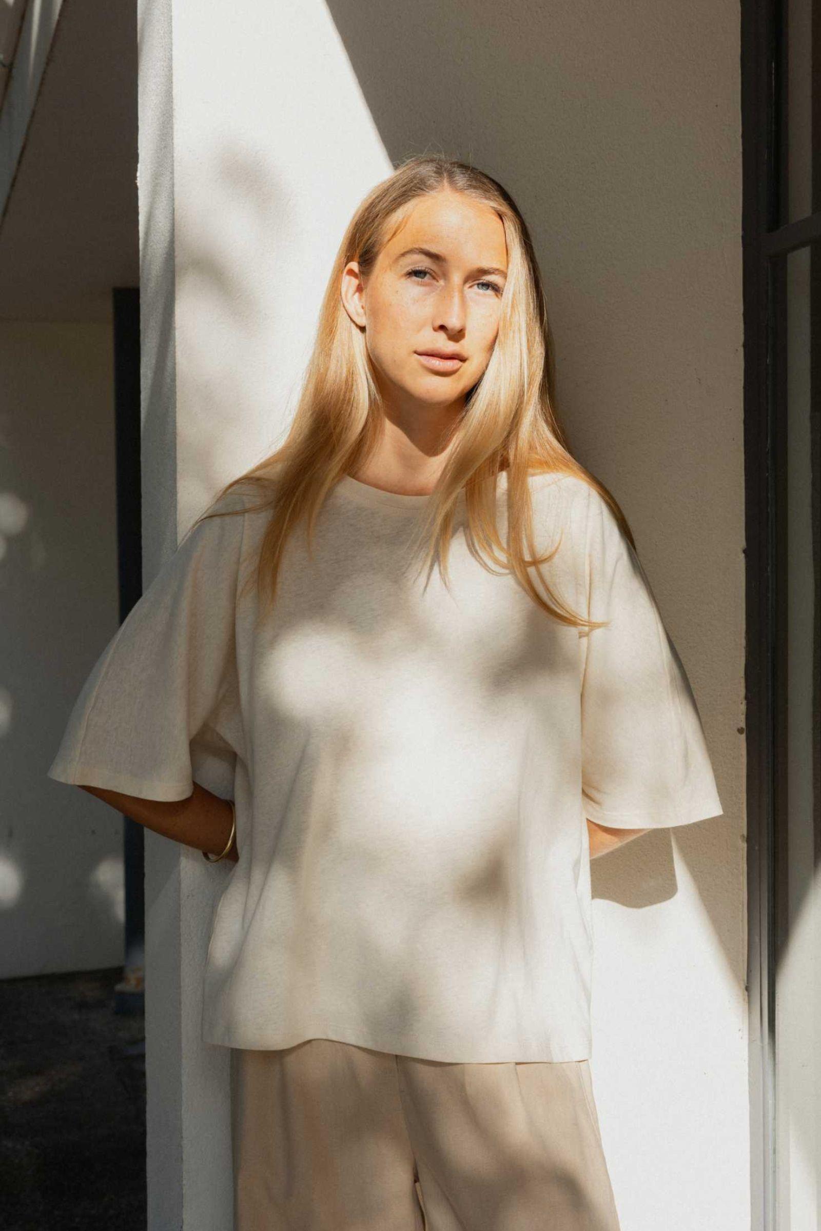 Woman wearing a white top and beige pants standing against a light-colored wall.