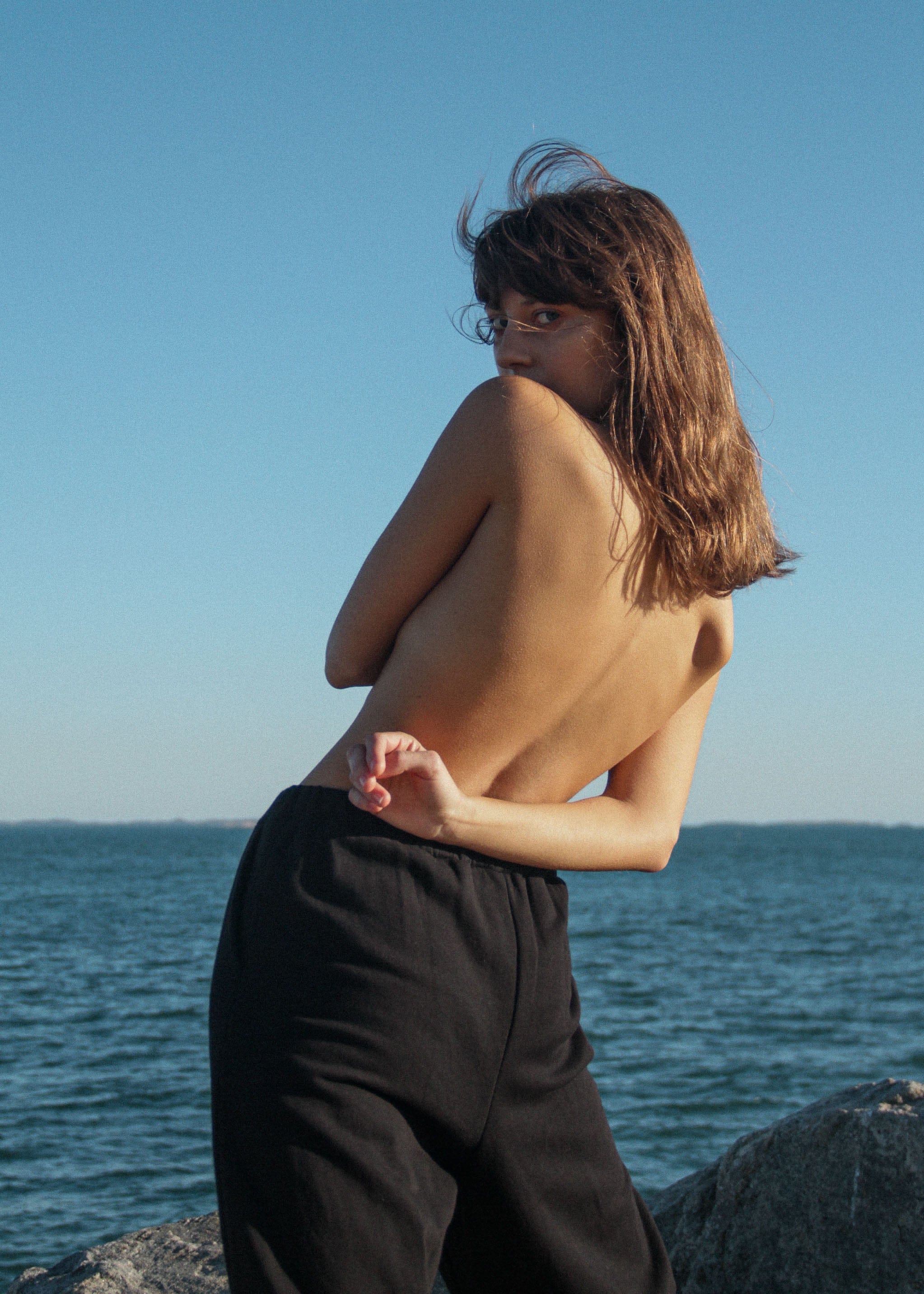 girl twists her torso towards the camera, she is at the seaside wearing black organic cotton trackpants.