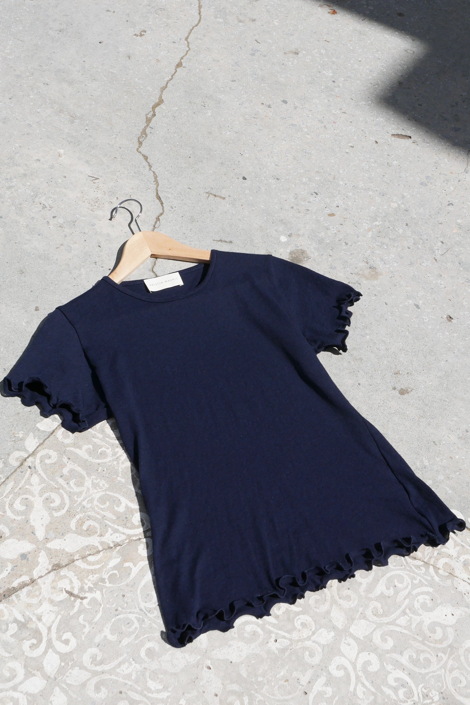 Navy blue ruffled tshirt on a hanger against a concrete floor.