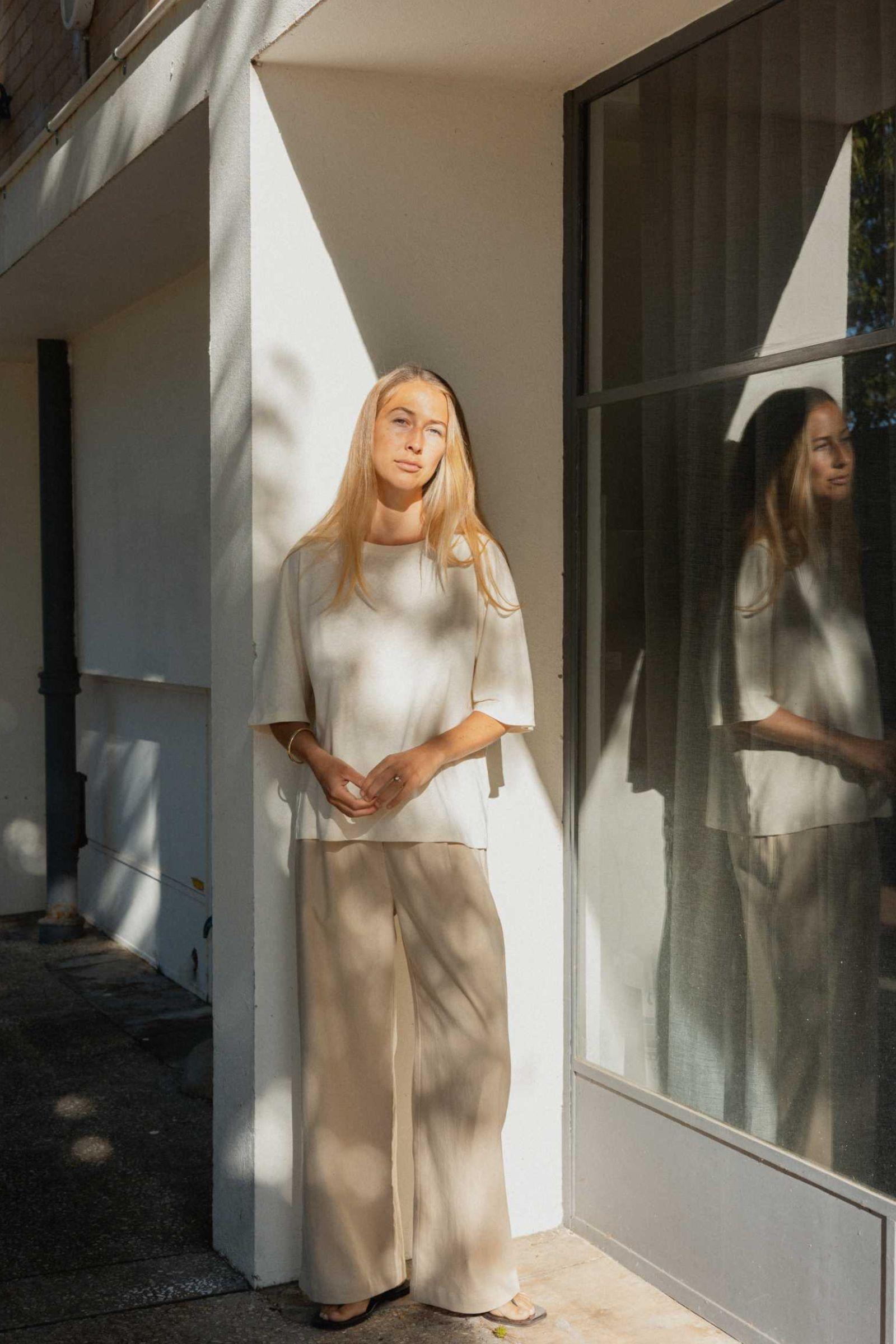 Woman standing in a sunlit area with a reflective surface behind her wearing cream refined tee